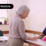 Idosa conversando com atendente enquanto assina um papel.
